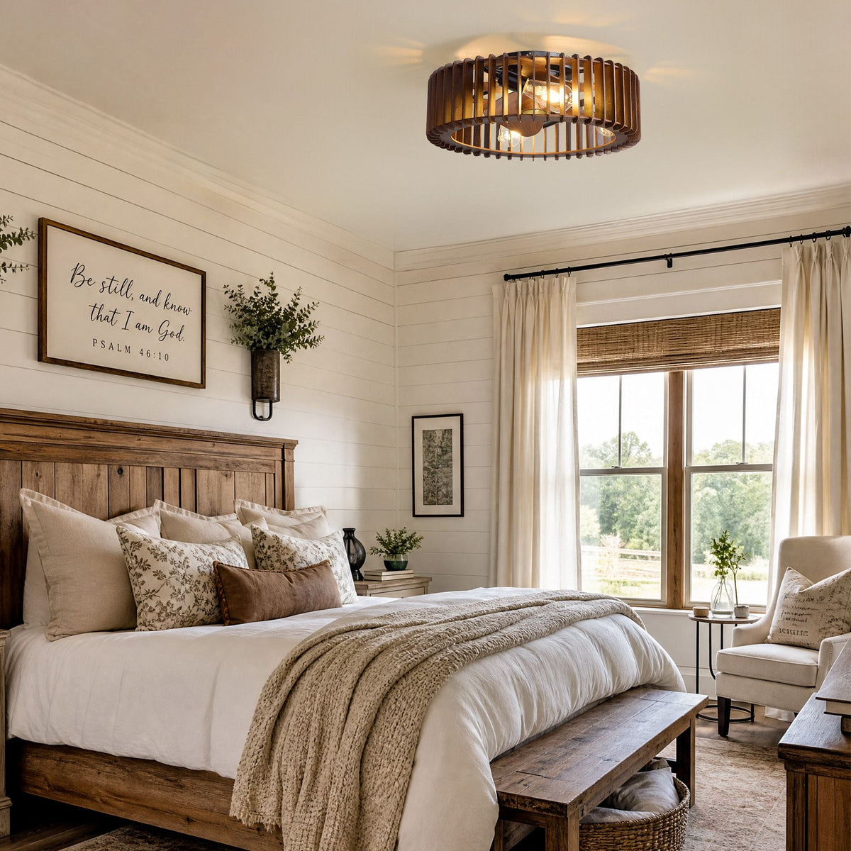 Industrial caged ceiling fan in farmhouse bedroom — dark wood bed, white bedding, decorative throw pillows, "Be still" wall art, large window with bamboo blinds and green views | CARRA HOME