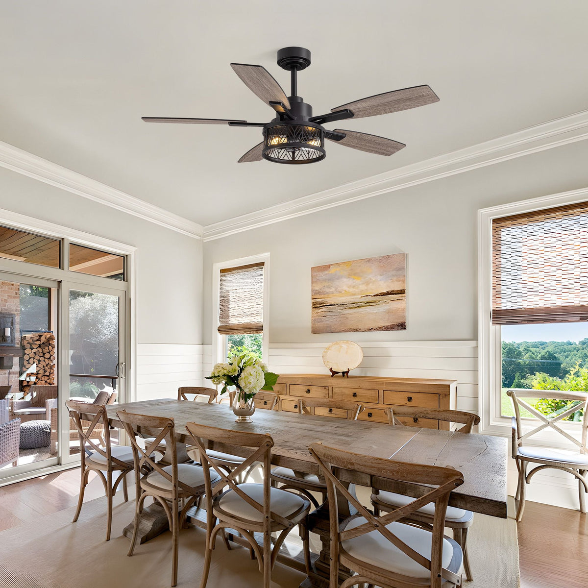 Farmhouse ceiling fan over dining area — long wood table, cross-back chairs, wicker shades, shiplap walls, outdoor view, rustic farmhouse style | CARRA HOME