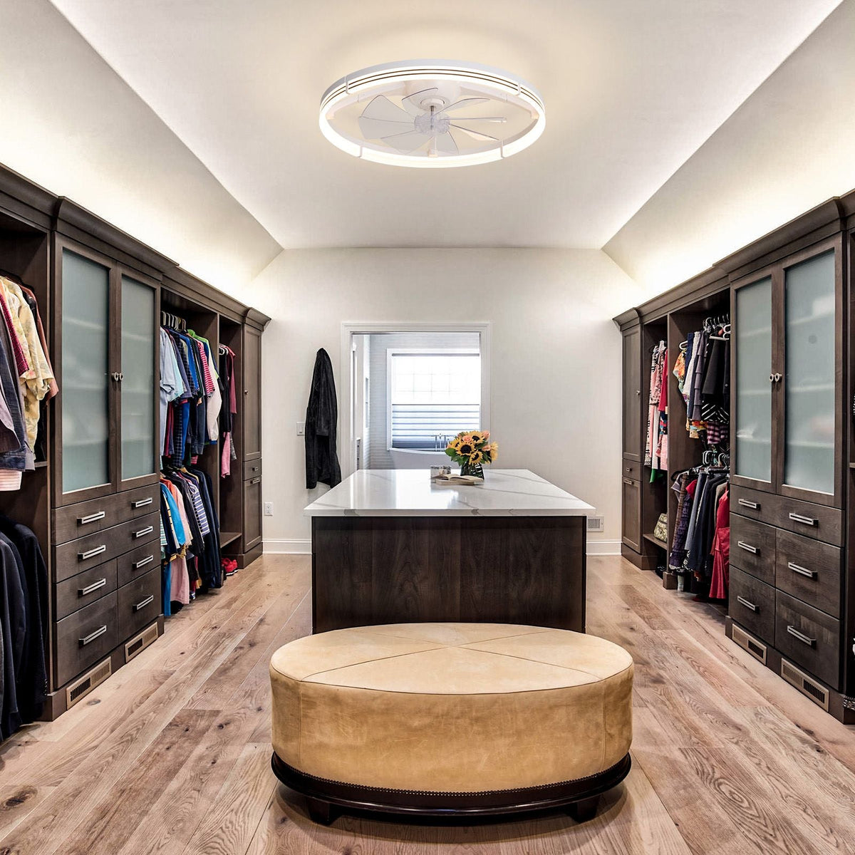 White foldable ceiling fan in luxury walk-in closet — dark wood cabinetry, frosted glass doors, white island with flowers, round ottoman, flush ceiling fan | CARRA HOME